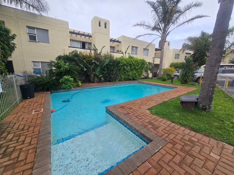 Communal swimming pool area featuring brick paving, established garden landscaping, and metal perimeter fencing within an apartment complex setting.