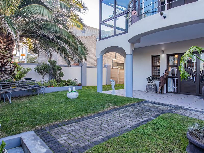 A well-maintained rear garden featuring a paved walkway, manicured lawn, and a covered patio area beneath a balcony with glass balustrades.