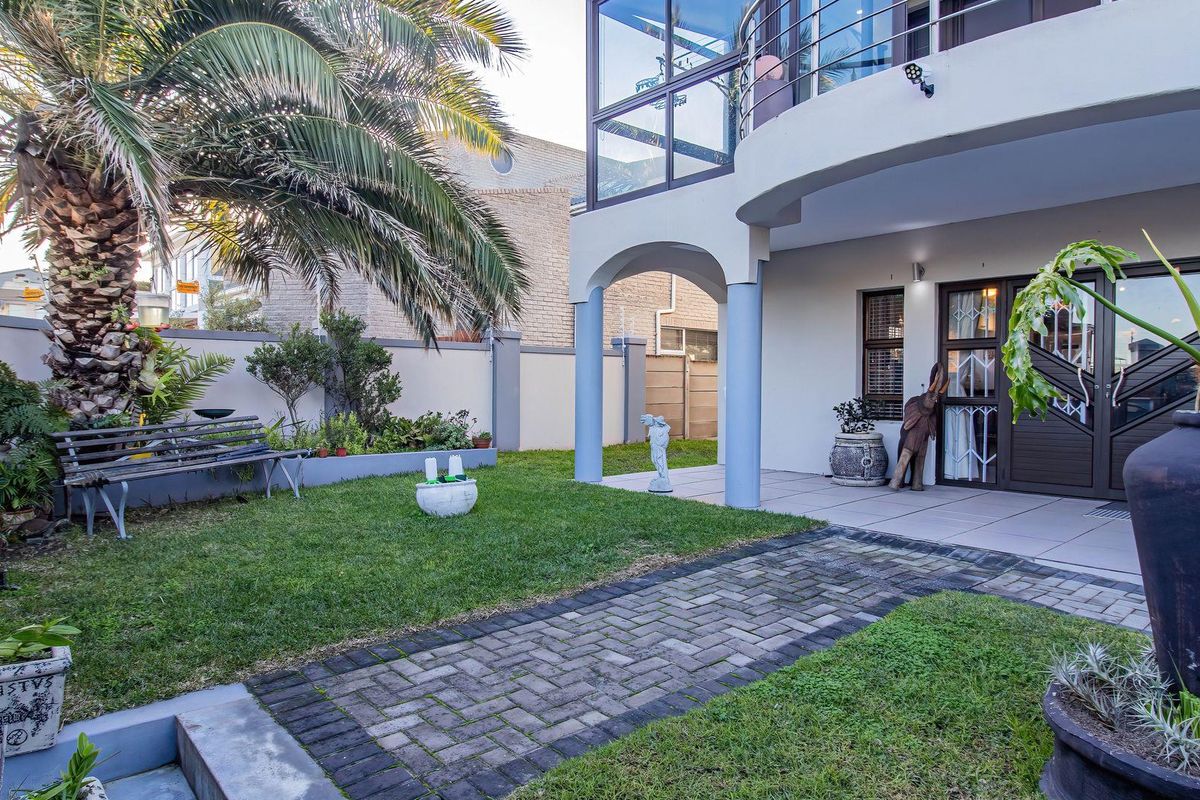 A well-maintained rear garden featuring a paved walkway, manicured lawn, and a covered patio area beneath a balcony with glass balustrades.