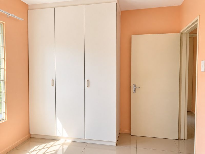 A bright bedroom featuring neutral tiled flooring, peach-toned walls, and a large white built-in wardrobe with ample storage space.