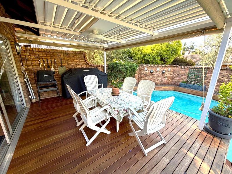 A covered timber deck patio area featuring a ceiling fan, overlooking a private swimming pool and enclosed by brick boundary walls.