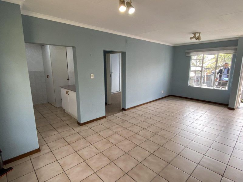 An open-plan living area featuring neutral ceramic floor tiles, light blue walls, and direct access to an adjoining kitchen and outdoor balcony space.