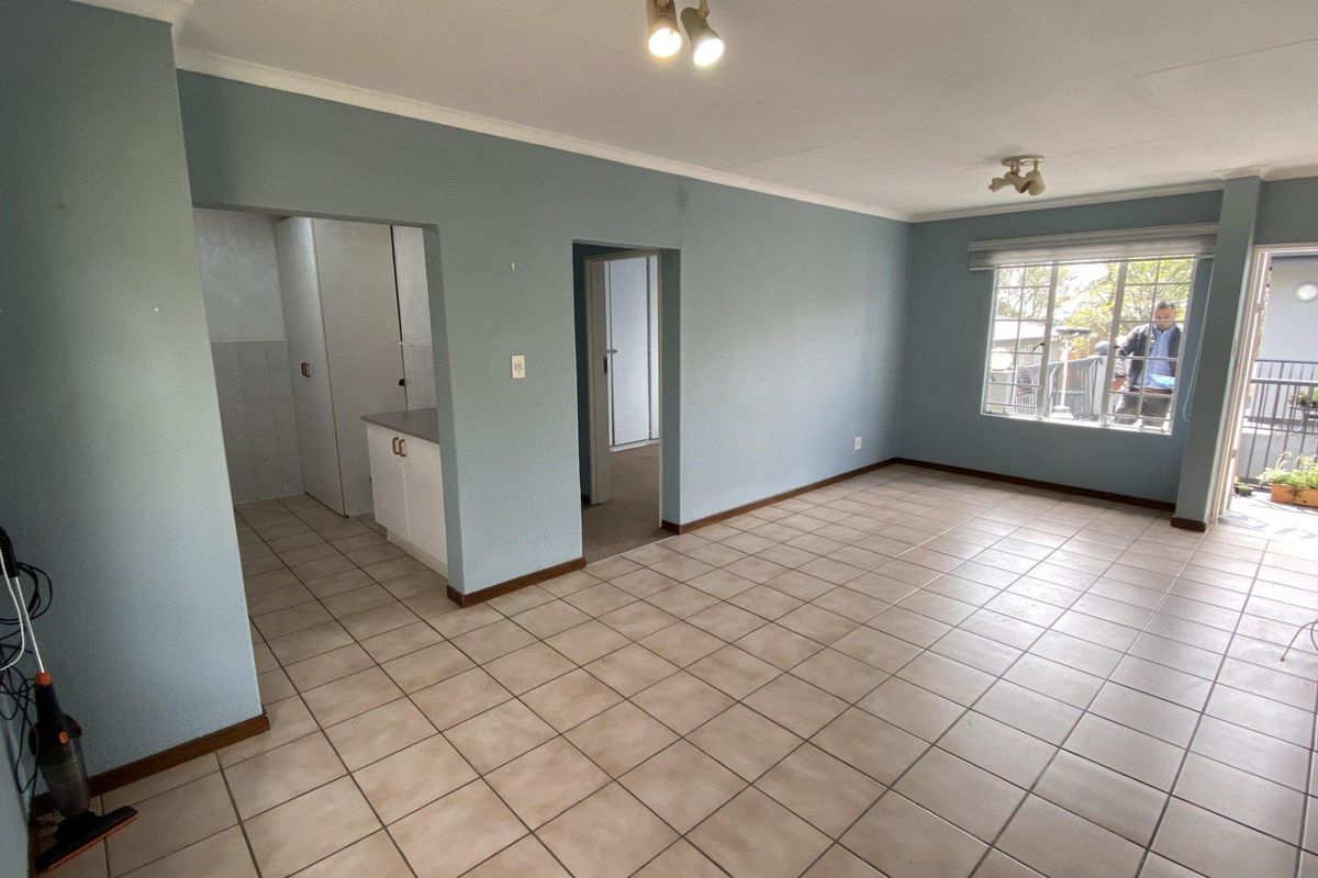 An open-plan living area featuring neutral ceramic floor tiles, light blue walls, and direct access to an adjoining kitchen and outdoor balcony space.
