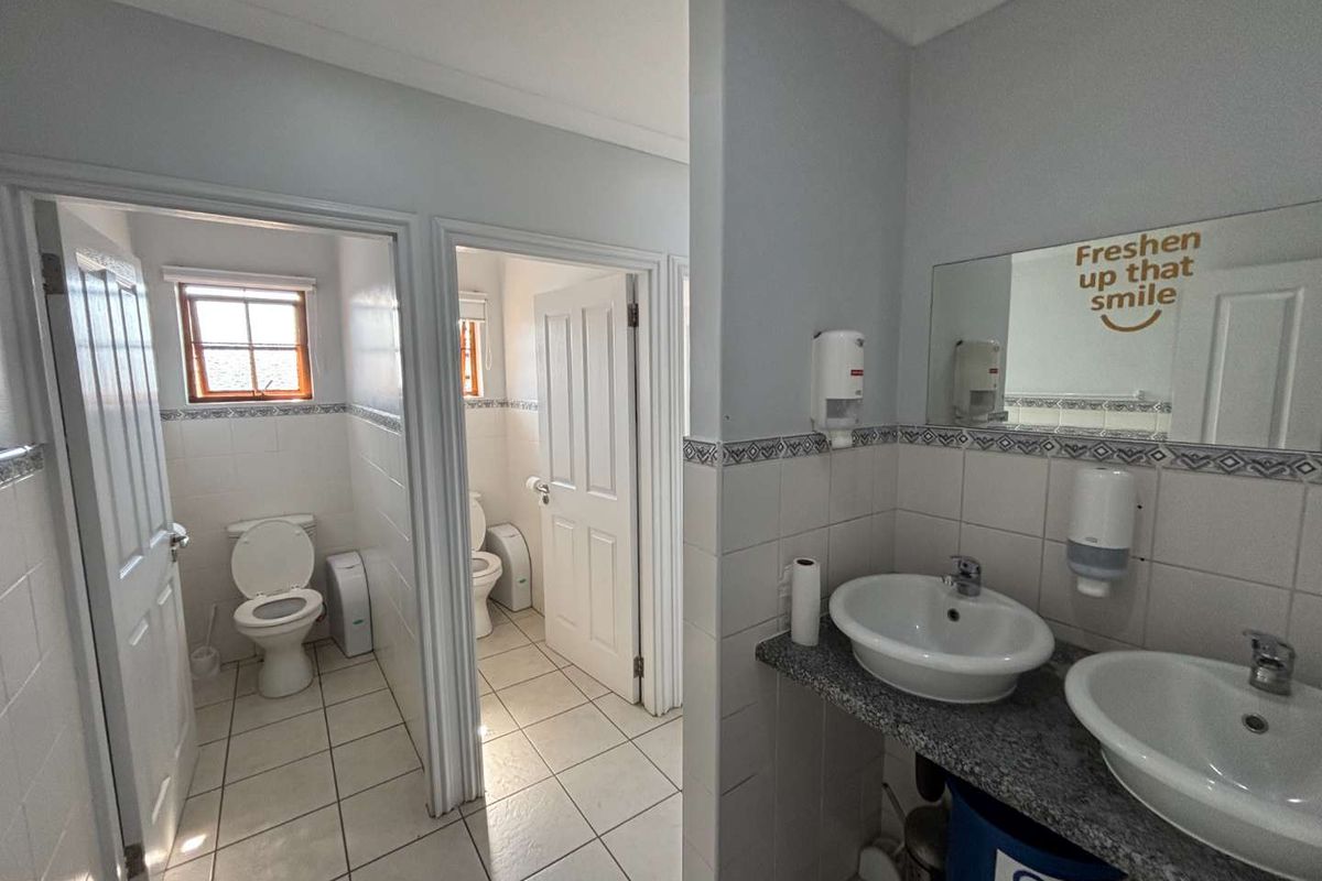 A functional office restroom facility featuring dual vanity basins, tiled walls, and separate toilet cubicles with natural light.