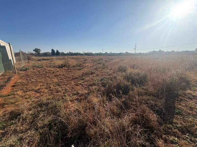 A wide-angle view of a vacant residential plot featuring dry grass, perimeter wire fencing, and clear sky.