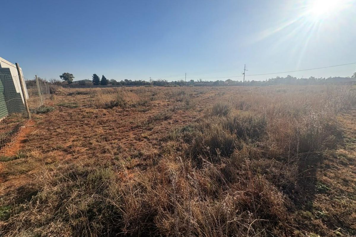 A wide-angle view of a vacant residential plot featuring dry grass, perimeter wire fencing, and clear sky.