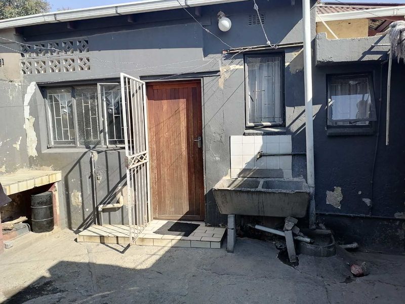 Rear exterior area featuring a concrete laundry trough, timber entrance door, and security gate, showing signs of significant wear and maintenance requirements.