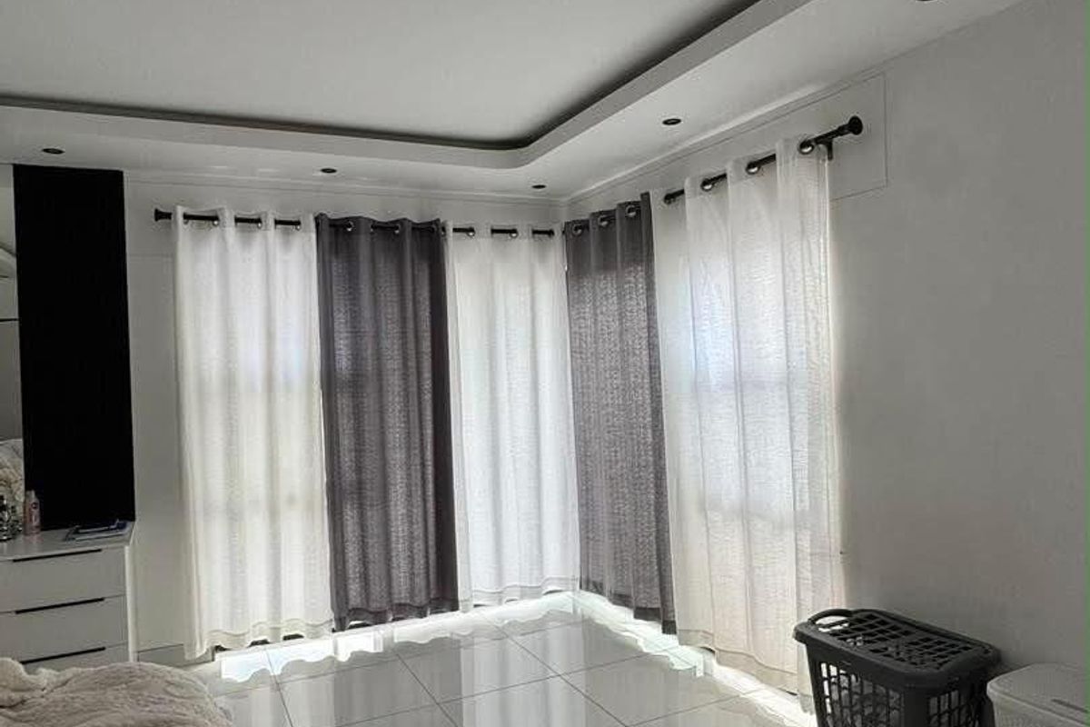 A bright bedroom featuring large windows with layered curtains, modern tiled flooring, and a recessed ceiling design with integrated lighting.