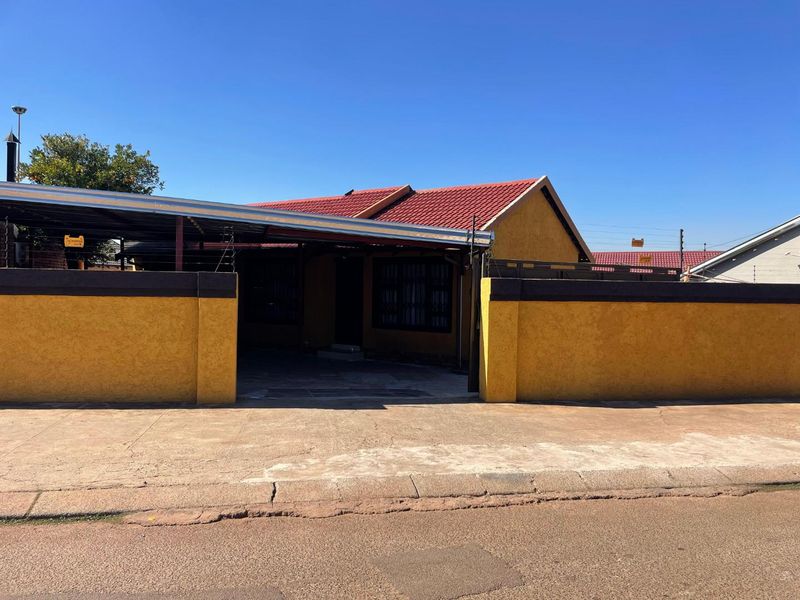Front view of a residential property featuring a yellow perimeter wall, a covered carport, and a tiled roof with security electric fencing.