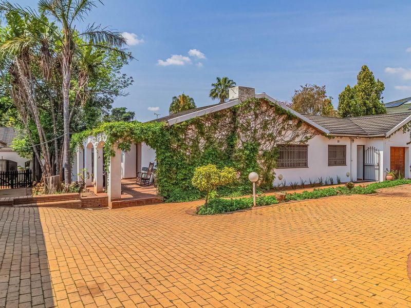 A charming front exterior featuring a spacious paved driveway, a covered patio area, and classic architectural details with established greenery.