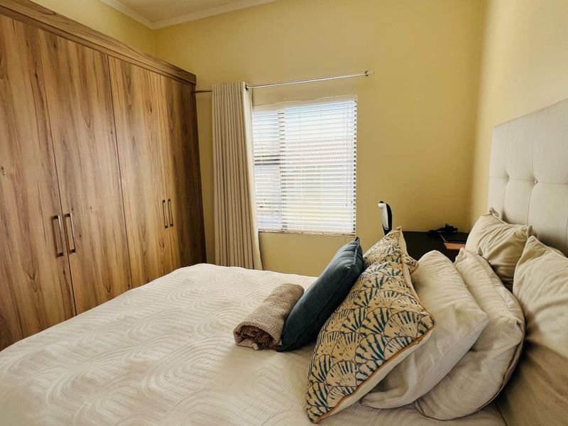 A well-maintained bedroom featuring extensive built-in wooden cupboards, neutral wall tones, and functional window treatments including venetian blinds and curtains.