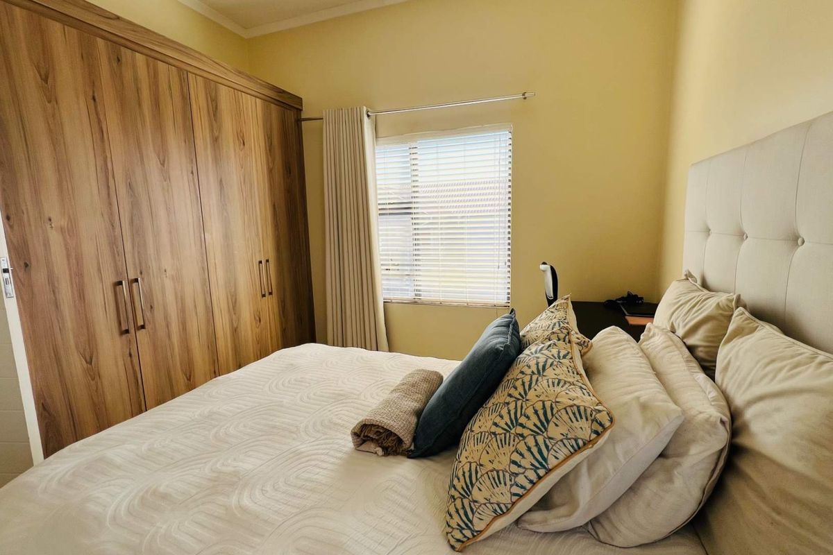 A well-maintained bedroom featuring extensive built-in wooden cupboards, neutral wall tones, and functional window treatments including venetian blinds and curtains.