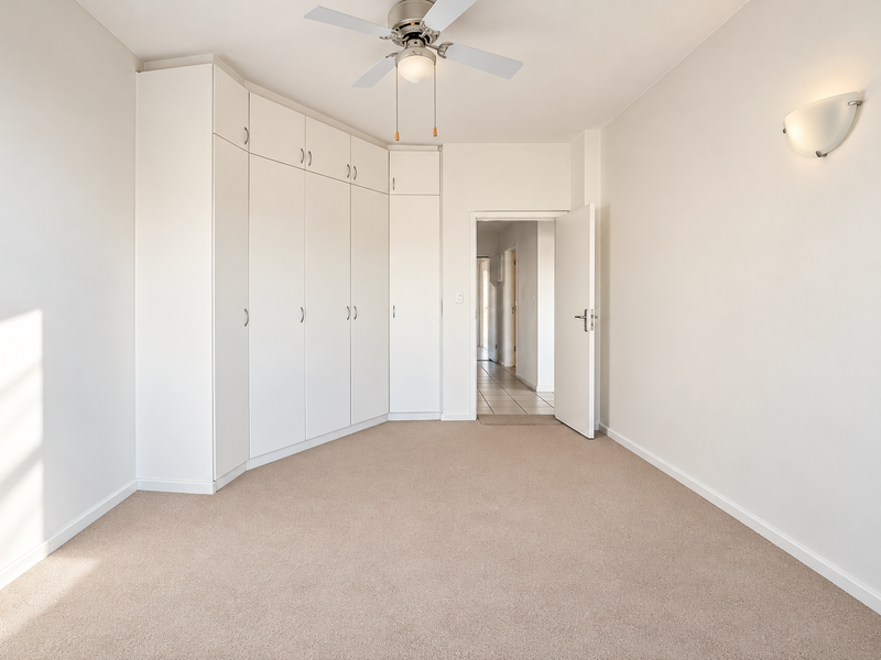 A bright, empty bedroom featuring neutral carpeting, ample built-in cupboard storage, a ceiling fan, and a wall-mounted light fixture.