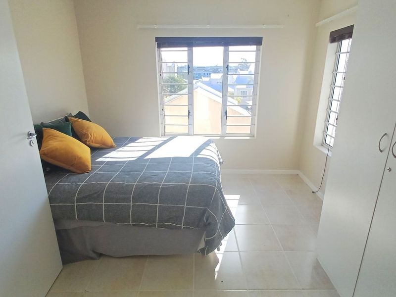 A functional bedroom featuring tiled flooring, built-in cupboards, and large windows with security bars, providing natural light and a view of the surrounding area.