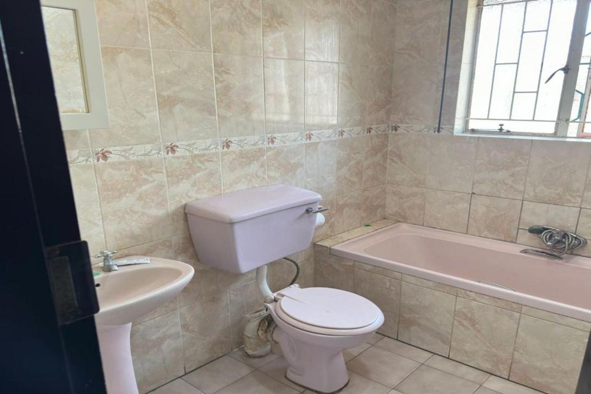 A dated bathroom featuring a pastel-colored bathtub, pedestal basin, and toilet, finished with neutral wall tiles and a window with security bars.