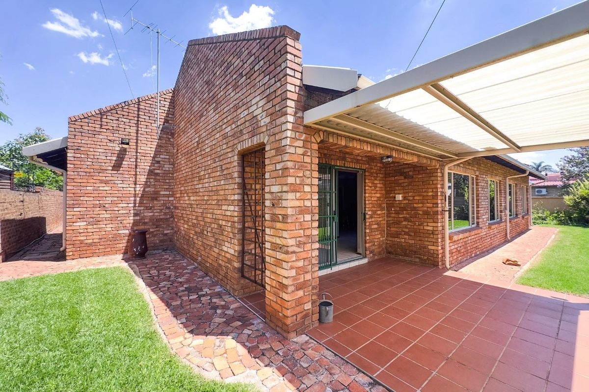 A rear view of the house featuring exposed face-brick walls, a covered patio area with tiled flooring, and a surrounding lawn.