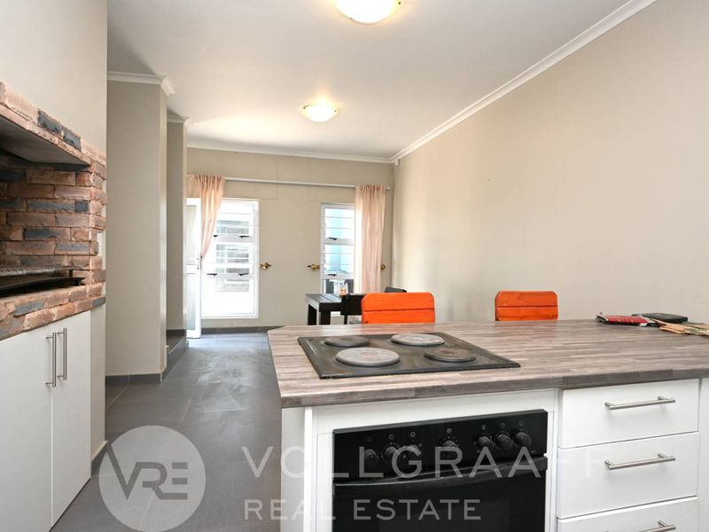 Open-plan living area featuring a kitchen island with built-in hob and oven, complemented by a rustic brick indoor braai and tiled flooring.