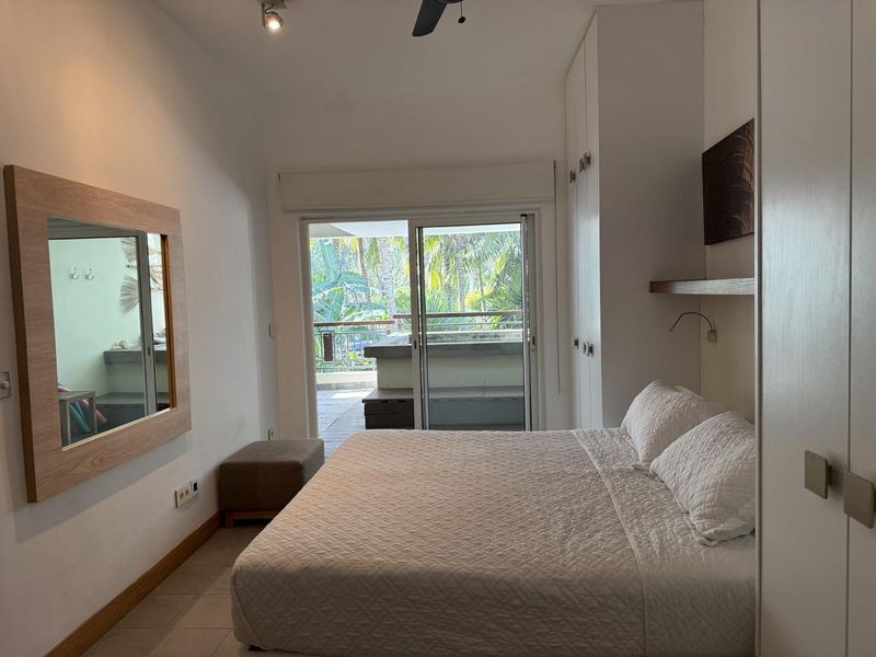 A well-maintained bedroom featuring built-in wardrobes, tiled flooring, and sliding glass doors that open onto a private balcony with lush greenery views.