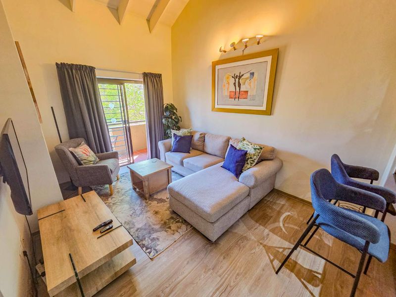 A bright living room featuring vaulted ceilings, laminate flooring, and a sliding glass door providing access to an outdoor balcony area.
