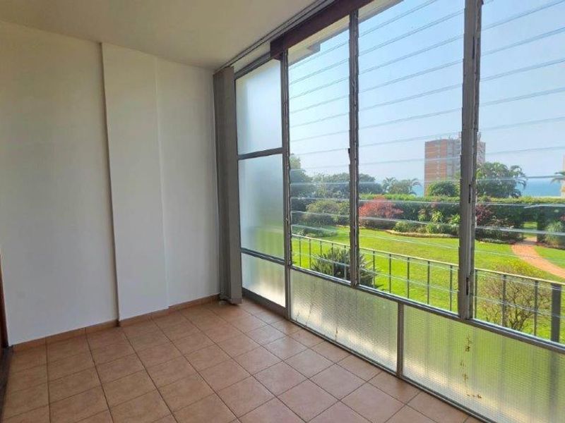 An enclosed balcony featuring tiled flooring, built-in storage, and expansive louvre windows offering pleasant garden and coastal views.