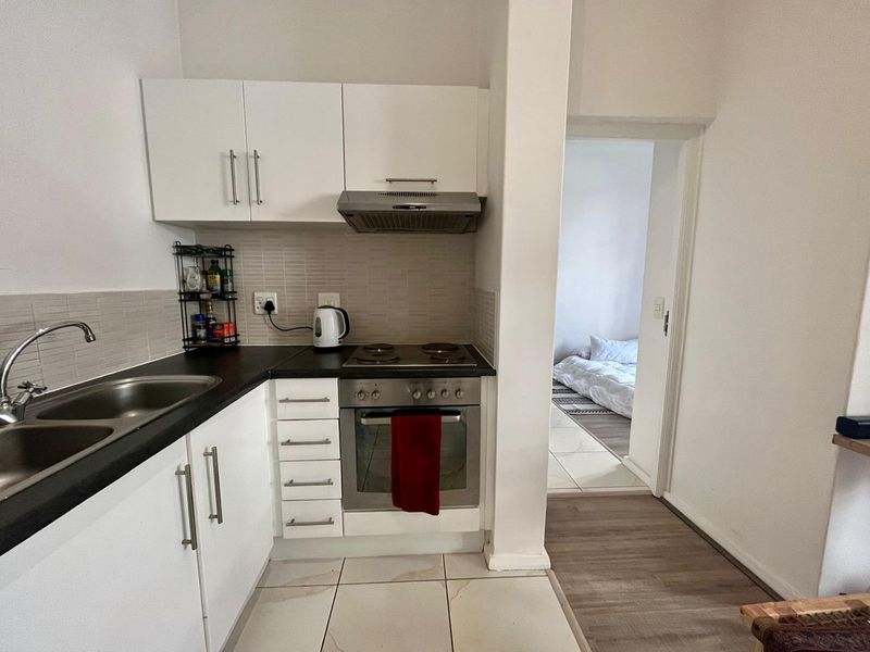 Compact kitchen featuring white cabinetry, dark countertops, integrated oven and hob, and tiled backsplash, with direct access to the adjacent bedroom.
