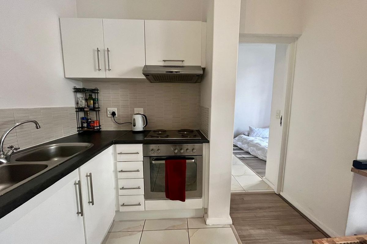 Compact kitchen featuring white cabinetry, dark countertops, integrated oven and hob, and tiled backsplash, with direct access to the adjacent bedroom.
