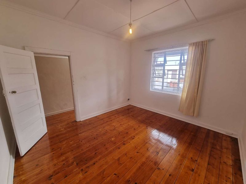 A bright, empty bedroom featuring polished hardwood flooring, a panel door, and a window fitted with security bars.