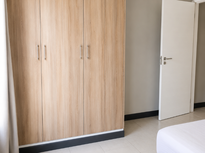 A clean bedroom featuring light wood-finish built-in cupboards, neutral wall tones, and tiled flooring.