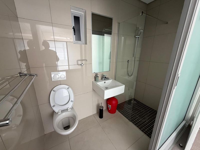 A modern, compact bathroom featuring a wall-mounted toilet, a wall-mounted basin, and a glass-enclosed shower with tiled finishes throughout.