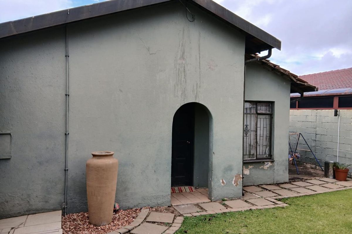 Rear exterior view of the house featuring an arched entrance, metal security bars on the window, and a paved walkway leading to the garden area.