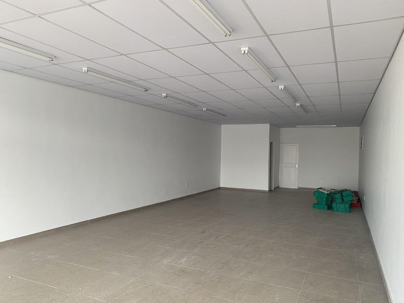 A spacious, empty retail unit featuring neutral tiled flooring, a suspended ceiling with integrated lighting, and clean white walls.