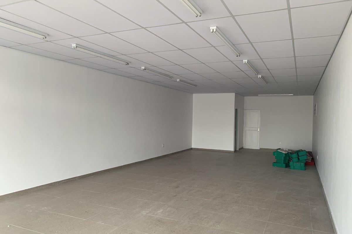 A spacious, empty retail unit featuring neutral tiled flooring, a suspended ceiling with integrated lighting, and clean white walls.