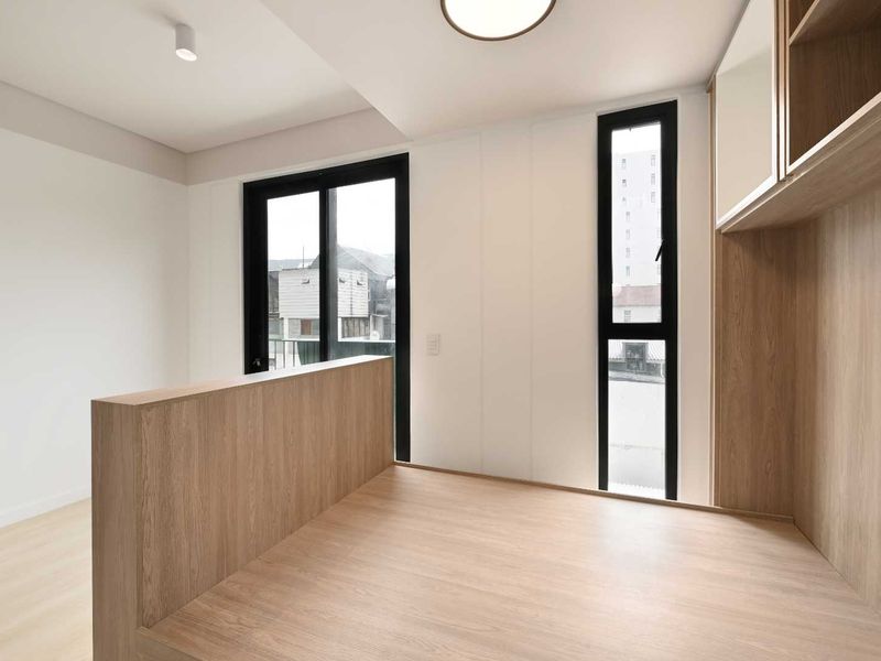 A modern, minimalist bedroom featuring a raised wooden platform, built-in cabinetry, and large black-framed windows providing natural light.