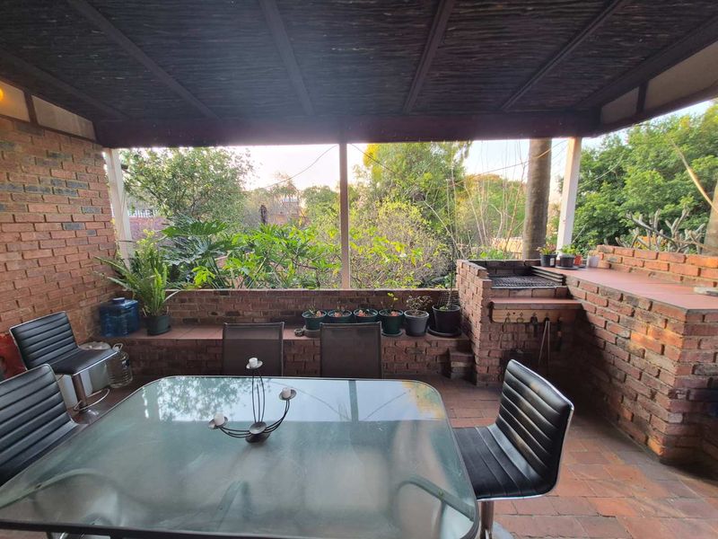 Covered outdoor patio featuring a built-in brick braai, brick paving, and a thatched roof, overlooking a garden area.