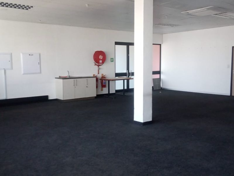 An open-plan commercial space featuring dark carpeting, structural columns, recessed lighting, and a small built-in kitchenette area with a sink.