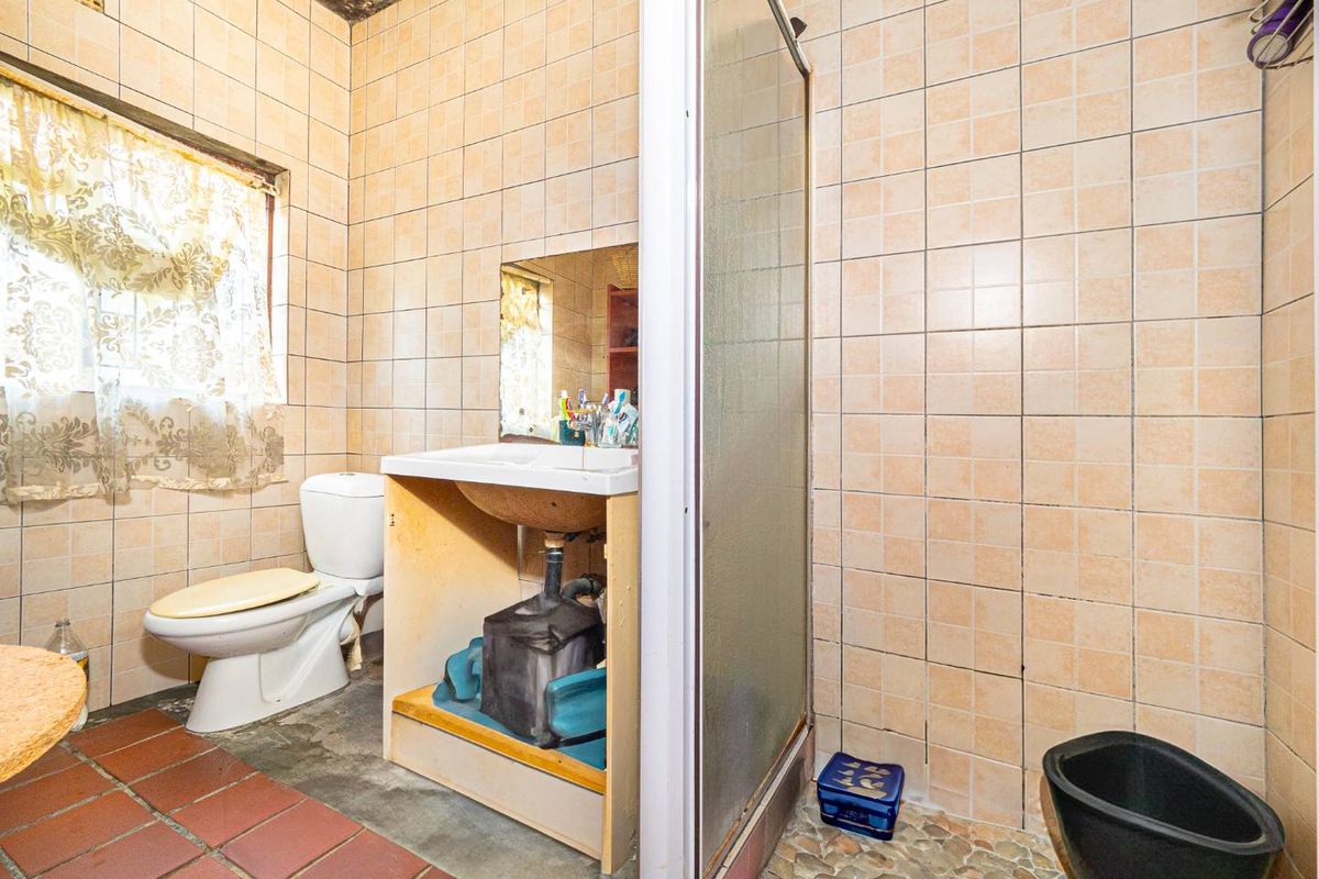 A dated bathroom featuring a shower enclosure, toilet, and a vanity basin with exposed plumbing and worn wall and floor tiling.