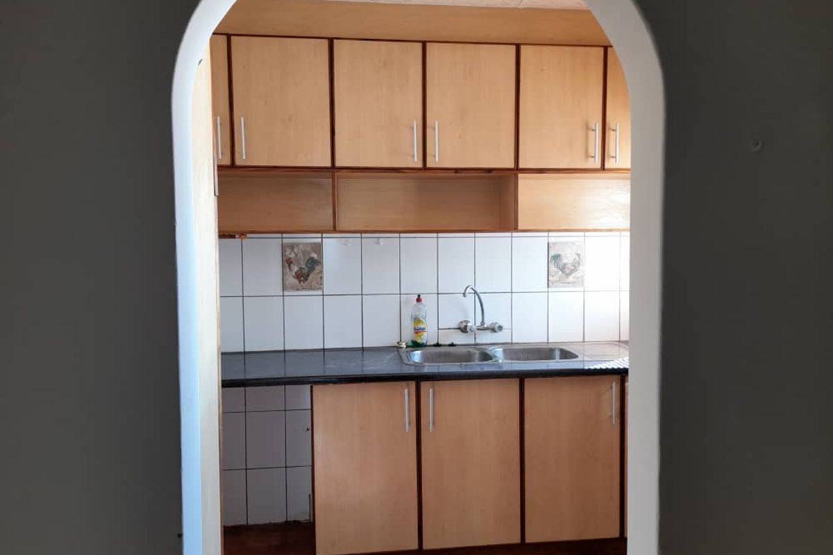 A dated kitchen featuring wooden cabinetry, a double basin sink, and tiled splashbacks, viewed through an arched doorway.