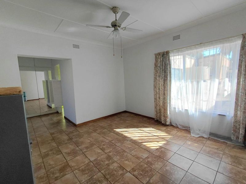 A bright, empty living room featuring ceramic floor tiles, a ceiling fan, and large windows with security bars, leading into a hallway with built-in cupboards.