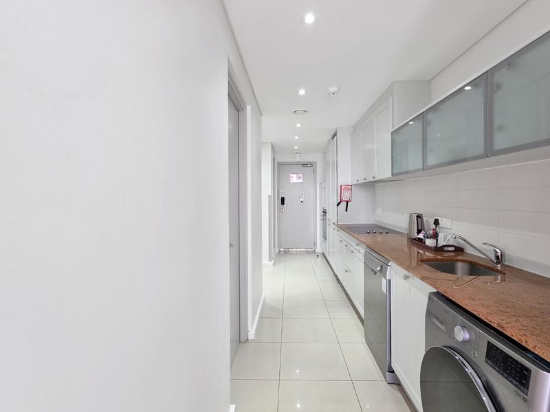 A modern galley-style kitchen featuring sleek white cabinetry, stone countertops, integrated appliances, and polished floor tiles in a bright, well-lit apartment space.