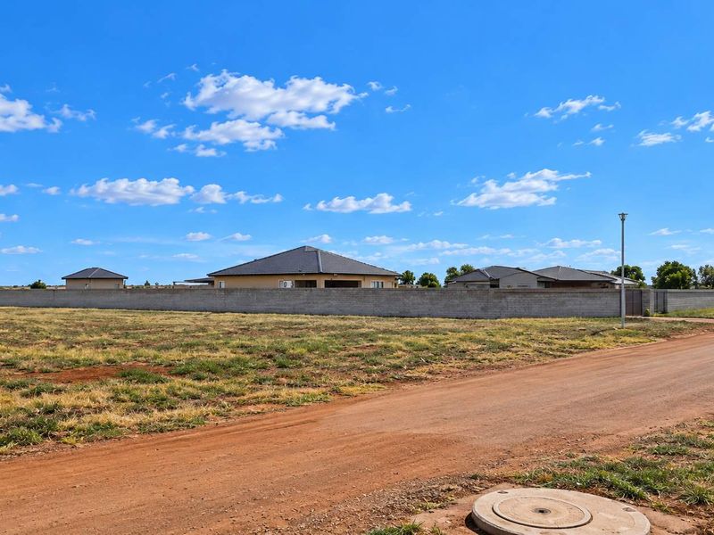 A view of a vacant residential plot featuring an unpaved road, perimeter wall, and nearby residential housing.