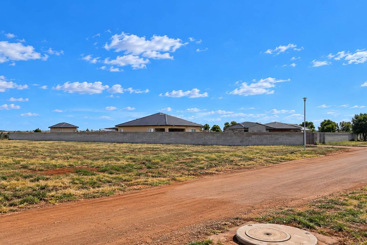 A view of a vacant residential plot featuring an unpaved road, perimeter wall, and nearby residential housing.