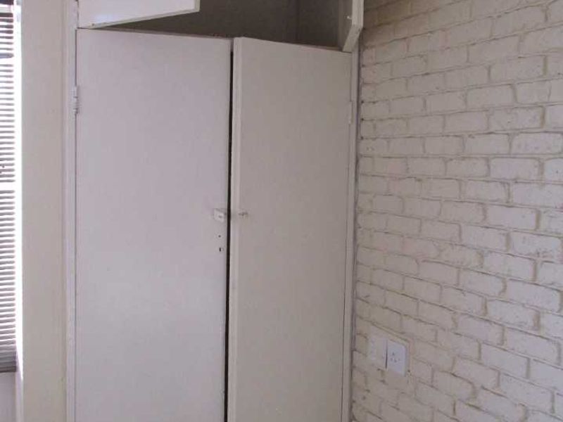 A bedroom interior featuring built-in cupboards with overhead storage and a painted brick feature wall, presenting a dated aesthetic in need of refurbishment.