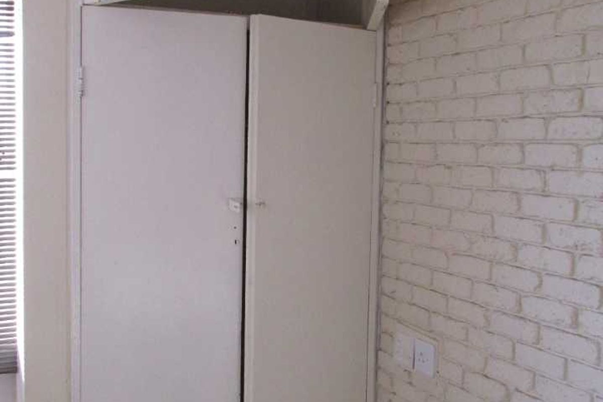 A bedroom interior featuring built-in cupboards with overhead storage and a painted brick feature wall, presenting a dated aesthetic in need of refurbishment.