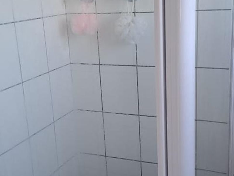A compact shower stall featuring white wall tiles and a glass enclosure, requiring some maintenance to the grout.