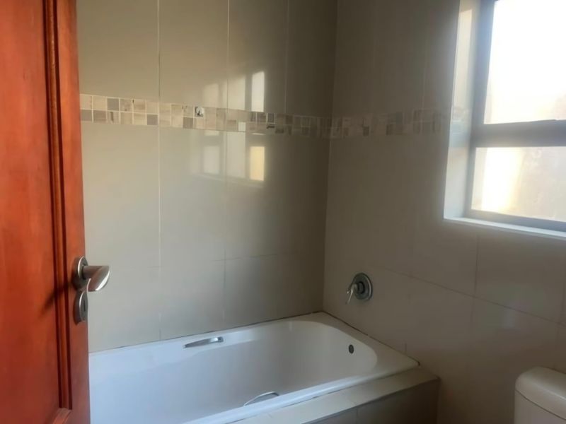 A functional bathroom featuring a built-in bathtub with wall-mounted mixer tap, neutral wall tiling with a decorative mosaic border, and natural light from a window.
