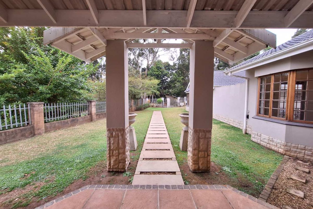 A rear garden view featuring a covered patio with stone-clad pillars, a timber pergola, and a paved walkway leading through the lawn.