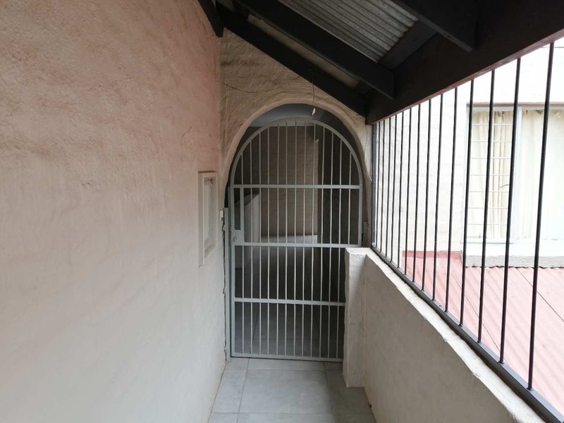 A covered balcony area featuring a security gate, metal balustrade, and tiled flooring, showing signs of wear on the wall finish.