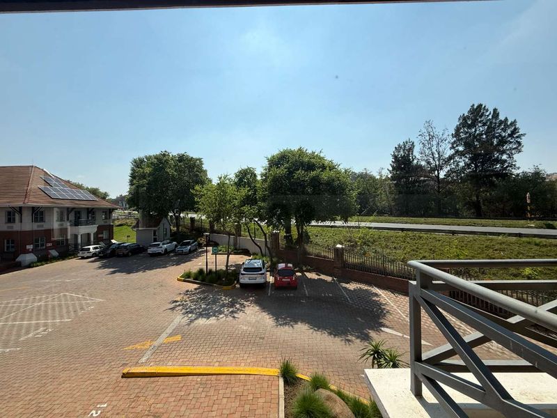 Elevated view overlooking a paved office parking area, surrounding greenery, and the adjacent building facade with solar panel installation.