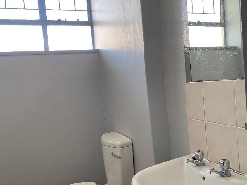 A functional bathroom featuring a pedestal basin, toilet, and tiled flooring, with natural light provided by a window with security bars.
