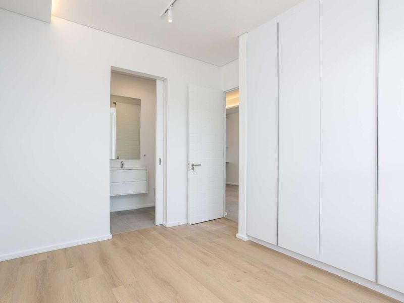 A bright, modern bedroom featuring light wood-look laminate flooring, ample built-in cupboard space, and direct access to an en-suite bathroom.